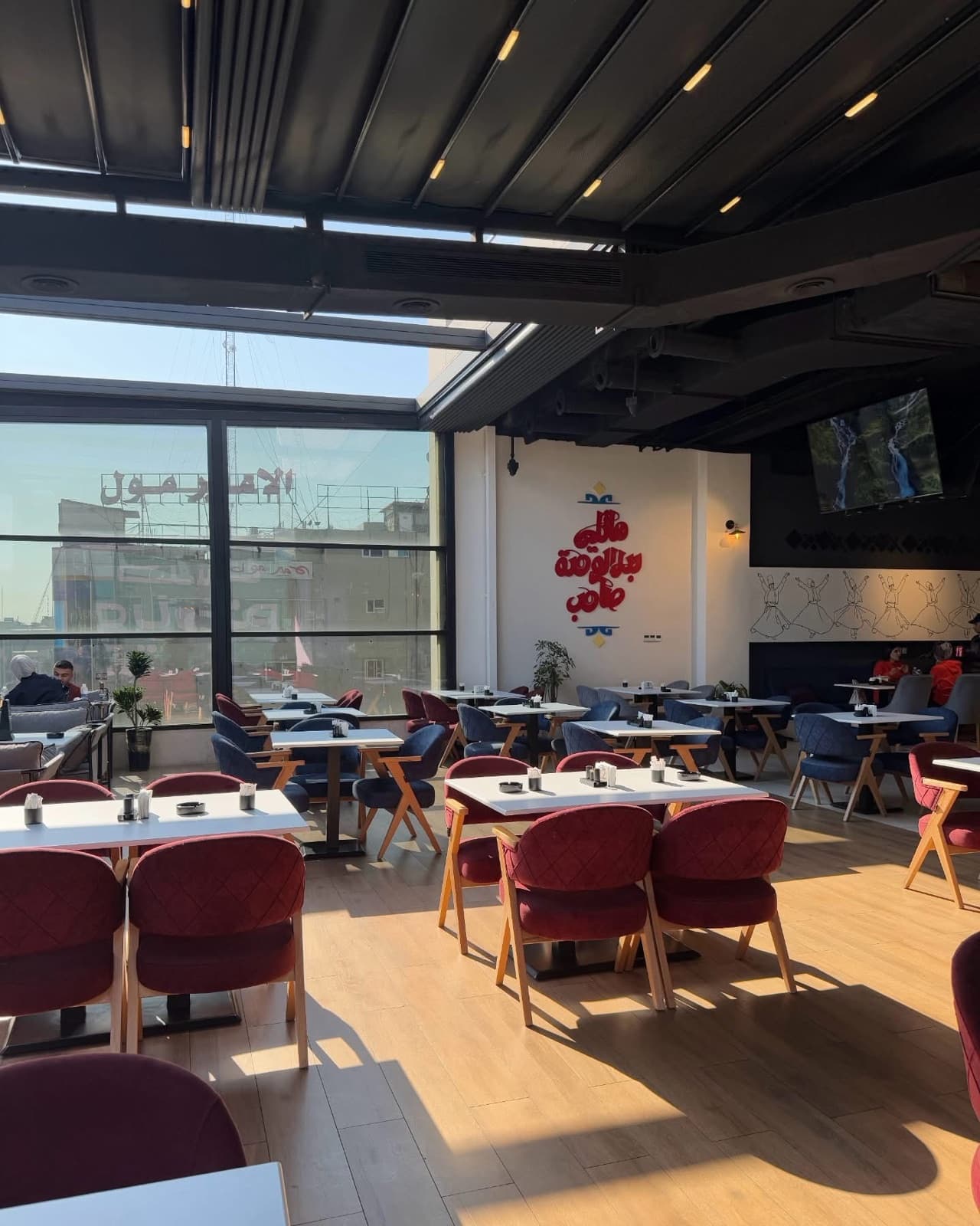 Sunlit dining interior with red and blue seating beneath a dark ceiling and large windows