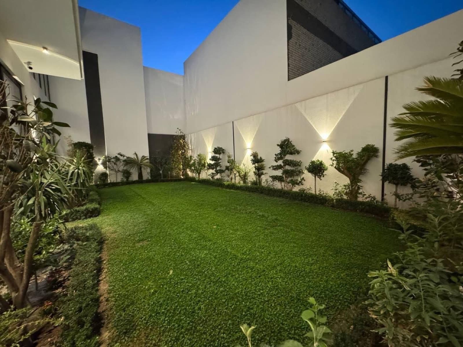 Landscaped courtyard with a central lawn, planted borders, and geometric wall lights at dusk