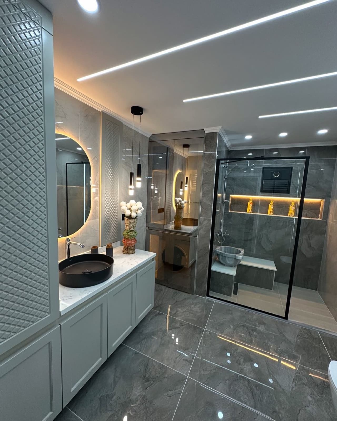 Grey-tiled bathroom with an illuminated round mirror, marble vanity, and glass shower enclosure