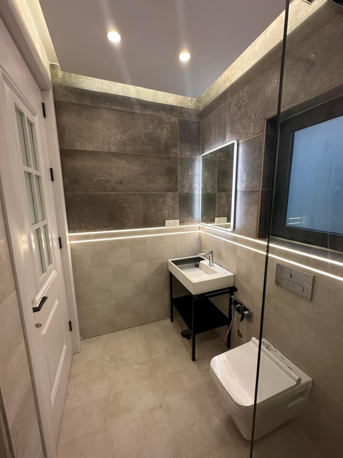 Compact bathroom with taupe wall tiles, an illuminated mirror, and a glass partition