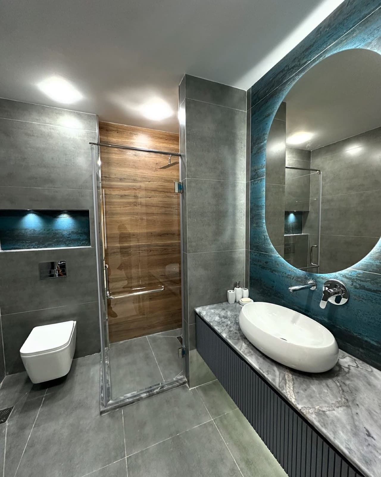 Grey bathroom with a blue-green backlit round mirror, marble vanity, and glass shower